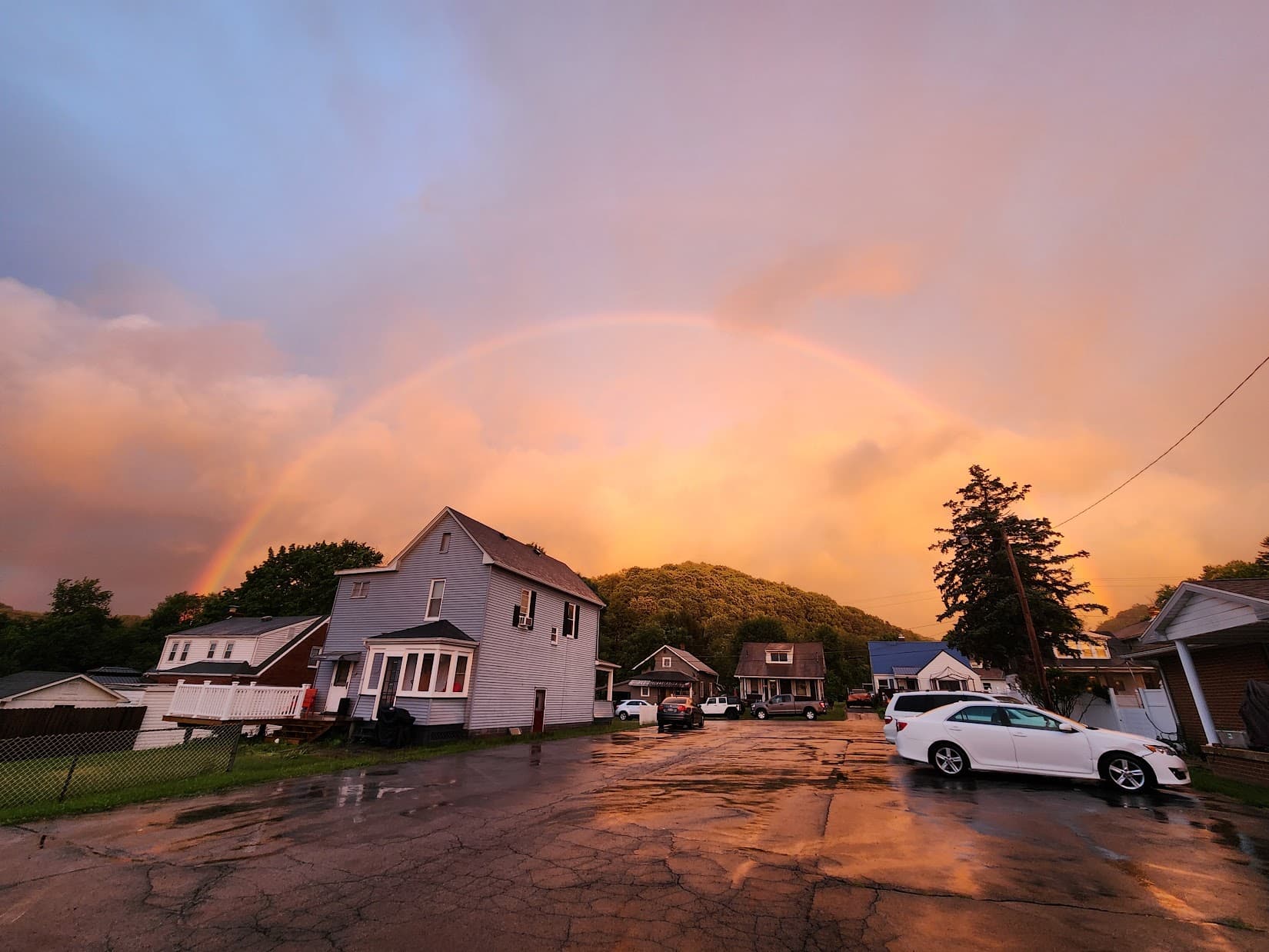 A rainbow over a wet street of houses in Chester, West Virginia, with a wooded hill behind them.