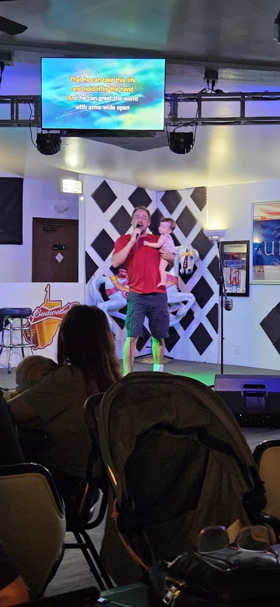 Tobe singing karaoke on stage while holding his baby granddaughter, with the lyrics on a screen above.